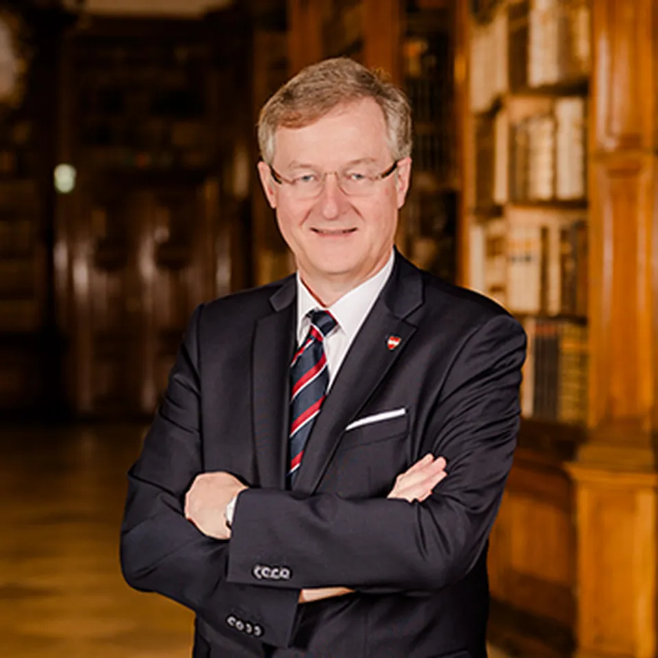 Ein Mann mit Brille, einem weißen Hemd mit rot-blauer Krawatte in einem dunklen Anzug.