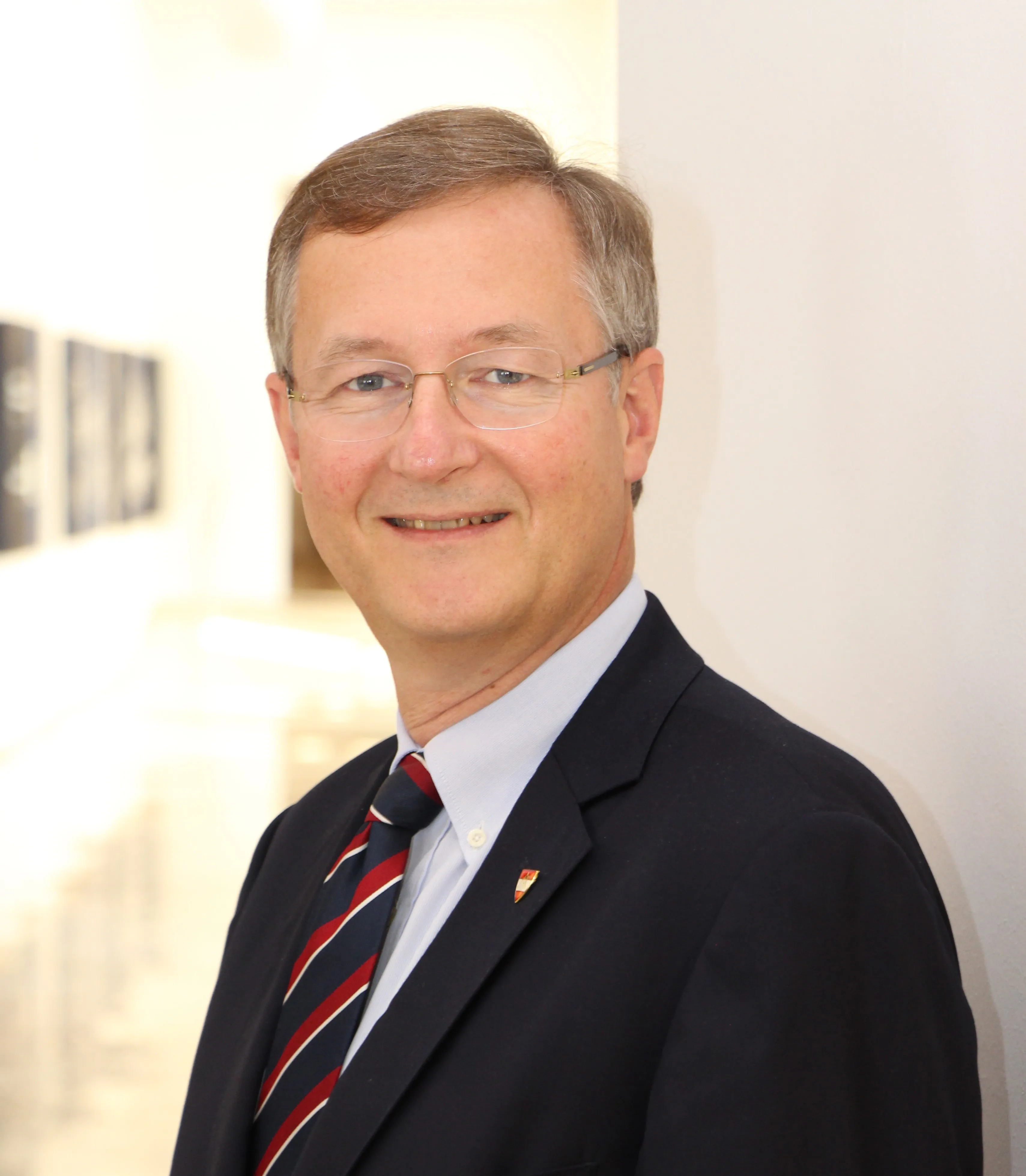 Ein Mann mit Brille, einem weißen Hemd mit rot-blauer Krawatte in einem dunklen Anzug.