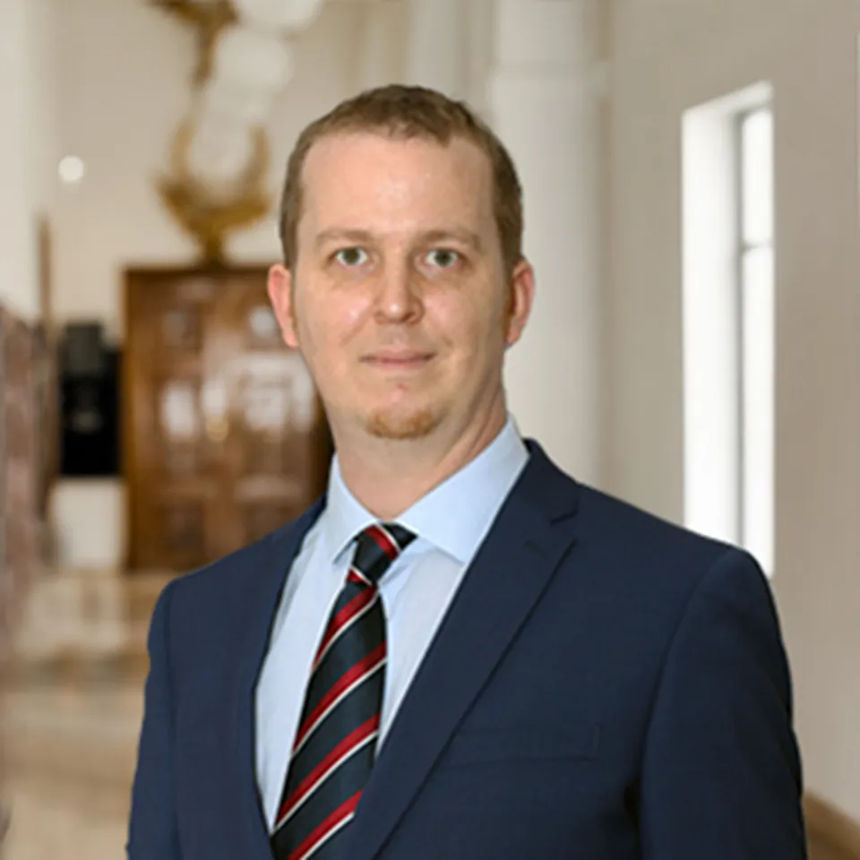 Mann mit kurzem Haar im dunkelblauen Anzug und rot-blauer Krawatte.