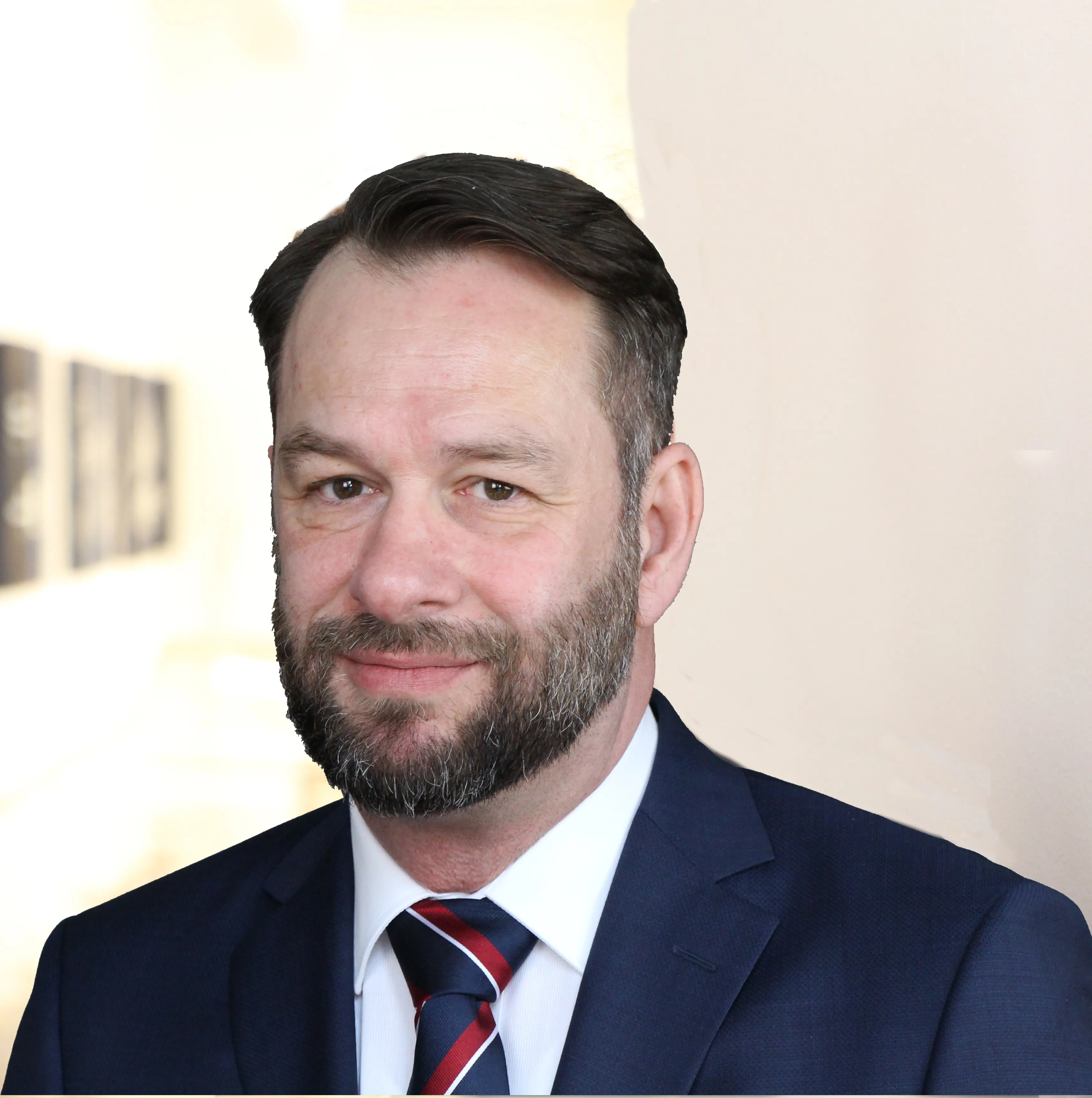 Mann mit kurzem dunklen Haar und Vollbart einem weißen Hemd mit rot-blauer Krawatte in einem dunklen Anzug.