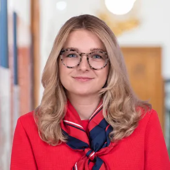 Portrait einer Frau mit blonden langen Haaren und Brille mit rot-blauem Seidentuch und roter Blazerjacke
