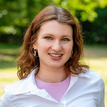 Portrait einer jungen Frau mit rotbraunen langen Haaren, rosa T-Shirt und weißer Bluse, in die Kamera lachend