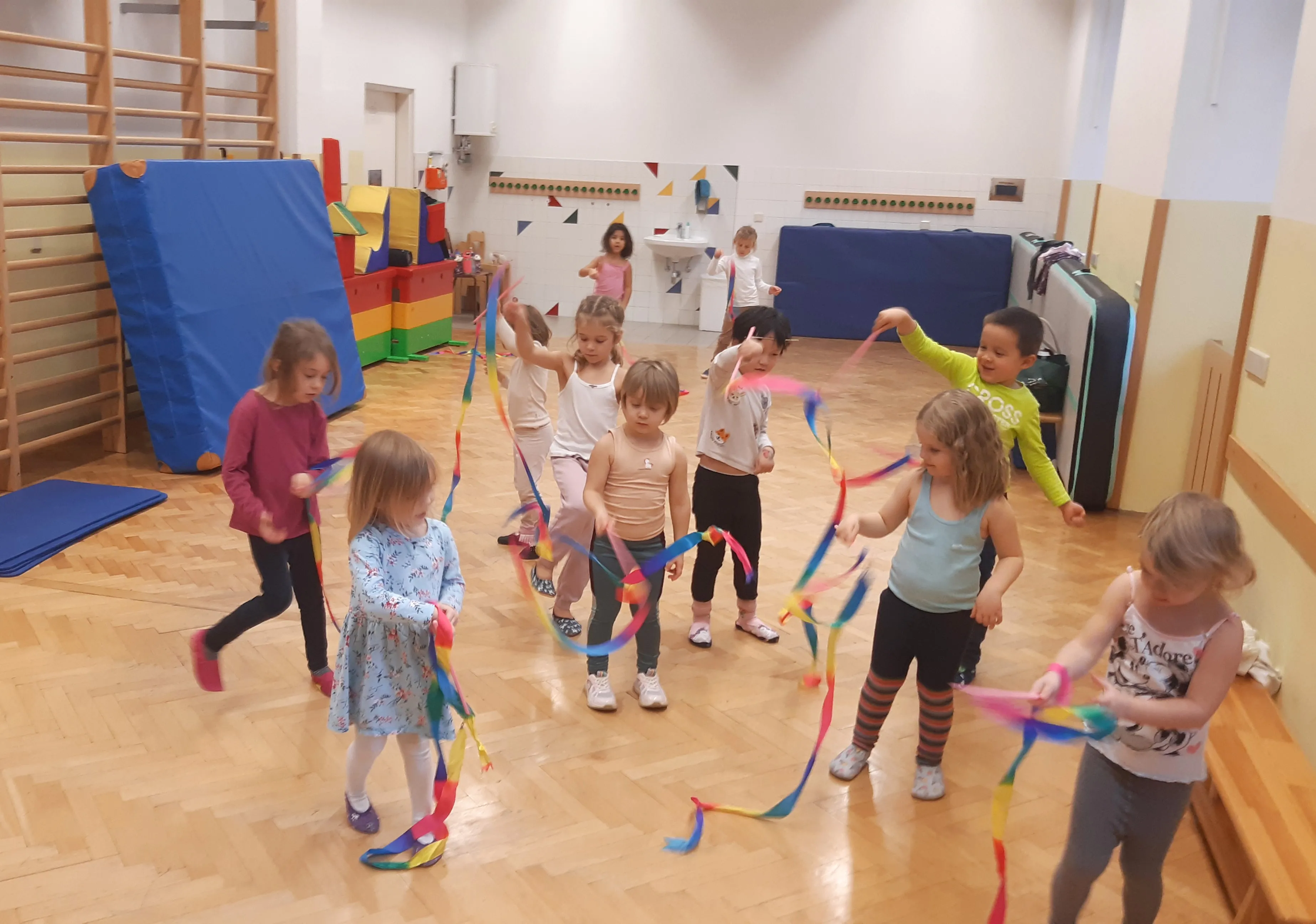 Gruppe von Kindern mit bunten Rhythmusbändern in einem kleinen Turnsaal mit Sprossenwand und bunten übergroßen Bauklötzen im Hintergrund