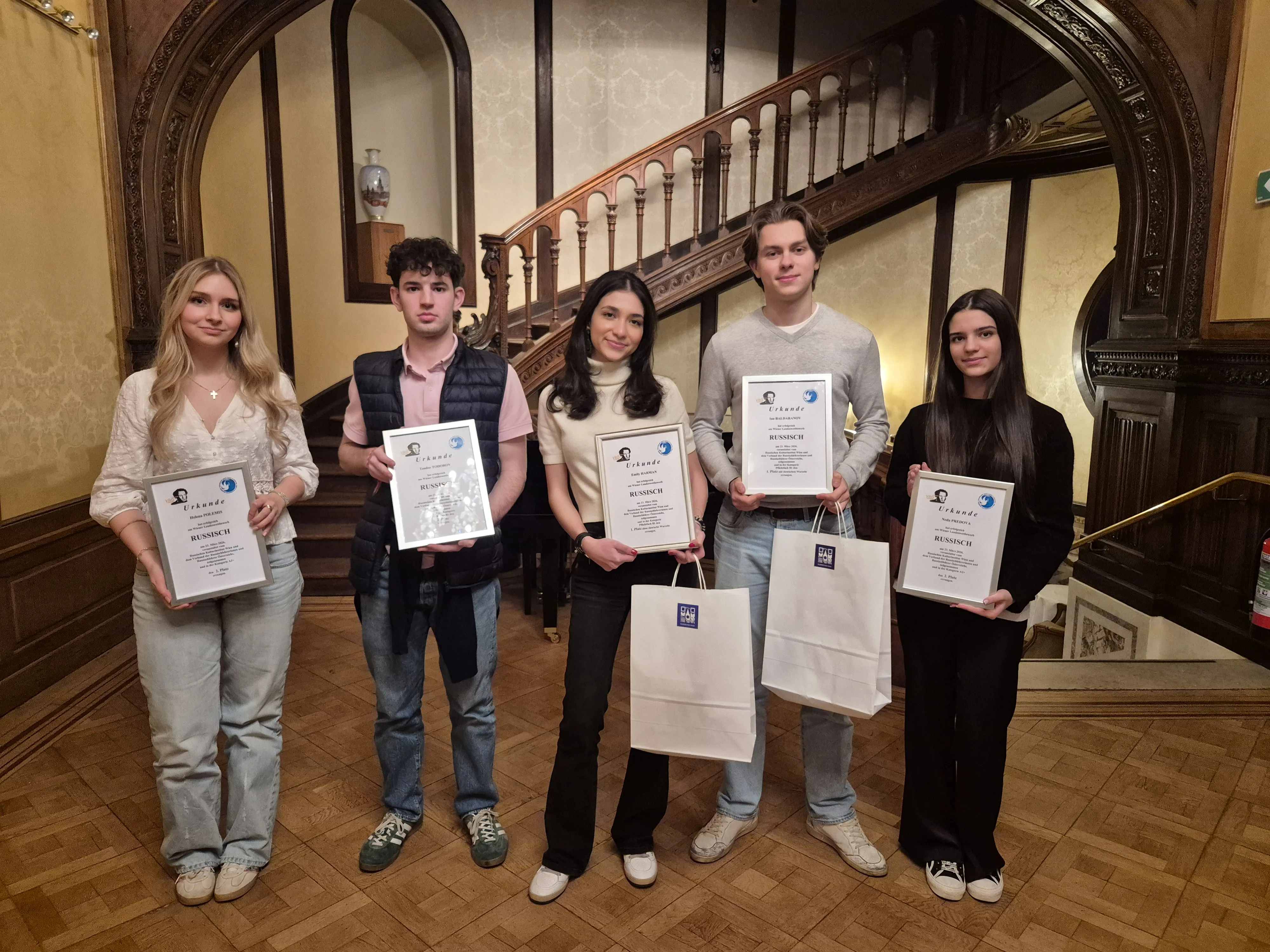 drei Mädchen und zwei Burschen zeigen stolz gerahmte Urkunden vor einem alten geschwungenem Treppengeländer