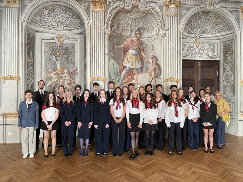 A group of people dressed formally stands together in an ornate room with elaborate frescoes on the walls and columns.