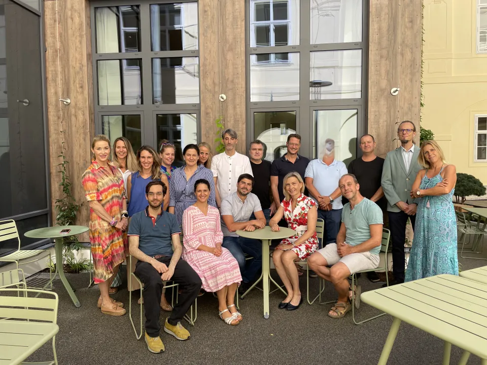 A group of people posing for a photo outside a building, with some sitting and others standing around a table with green chairs.