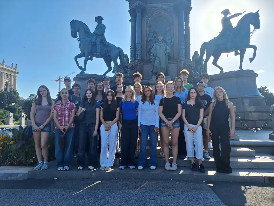 A group of young people posing in front of a monument with equestrian statues in a sunny outdoor setting.