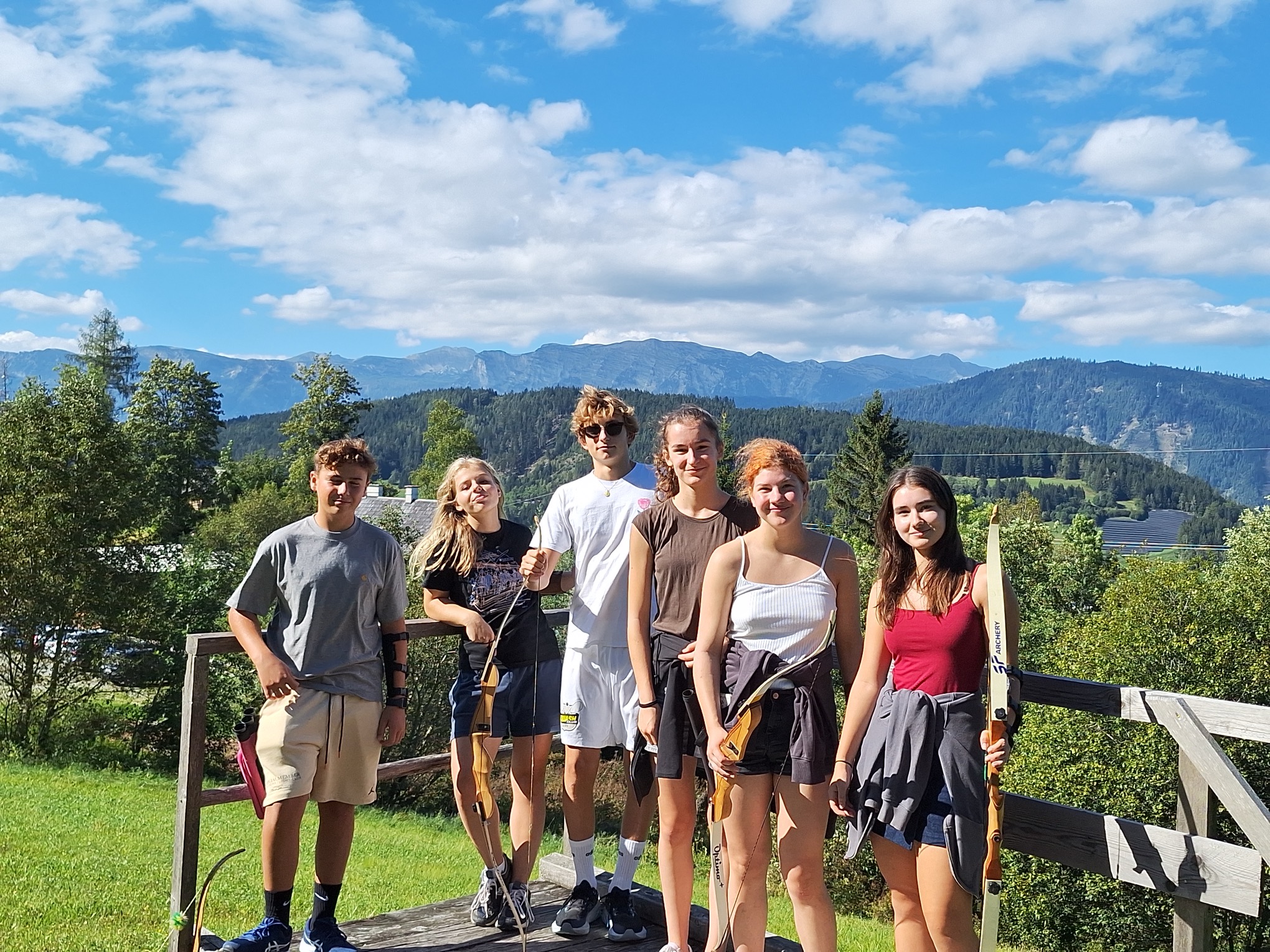 Gruppe von Teenagern mit vier Mädchen und zwei Burschen in lockerer Sommerkleidung auf einem Podest stehend jeweils einen Bogen in der Hand haltend vor prächtiger Natur- und Bergkulisse