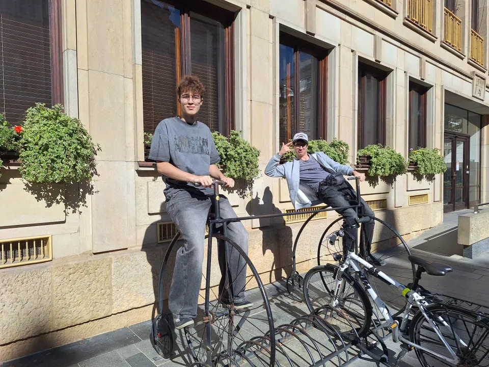Two people sitting on vintage penny-farthing bicycles outside a building with large windows and flower boxes.