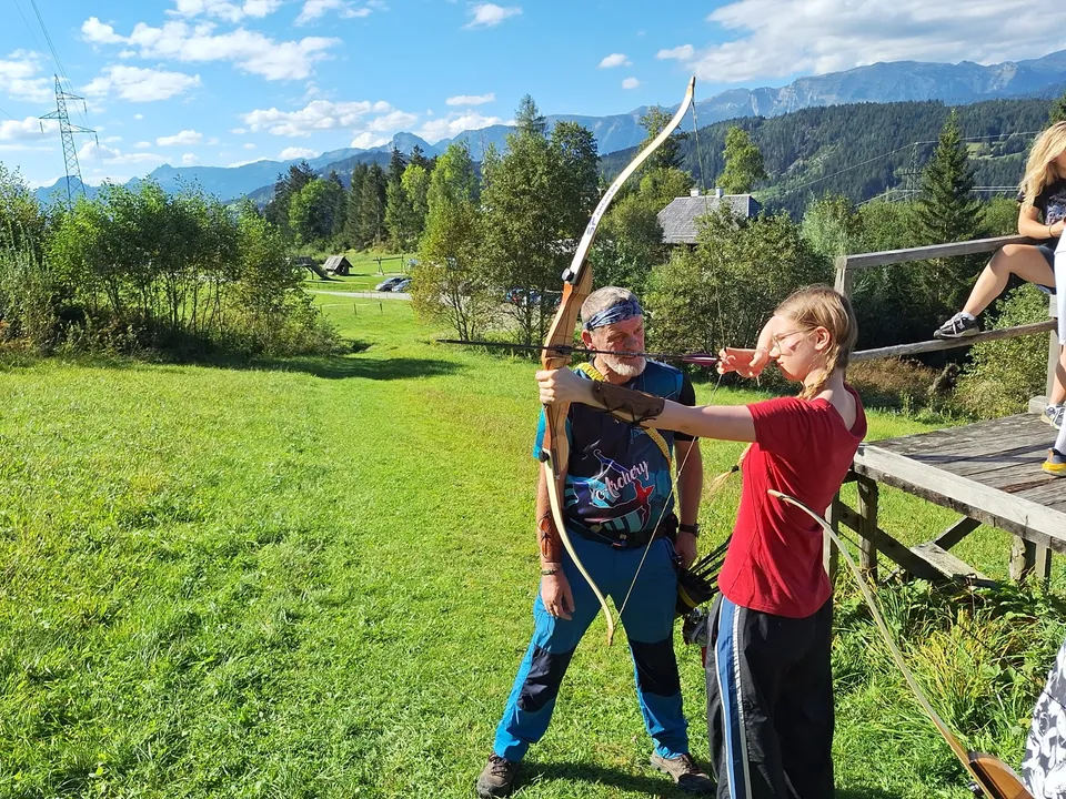 Eine junge Frau in rotem T-Shirt und blauer Jogginghose mit einem gespannten Bogen und ein Mann Instruktionen gebend in prächtiger Naturlandschaft.