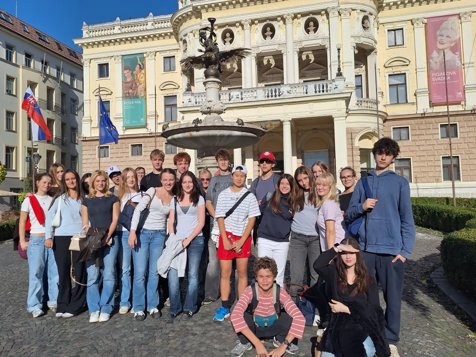 A group of people posing in front of an ornate building with banners, a fountain, and flags in the background, under a clear blue sky.