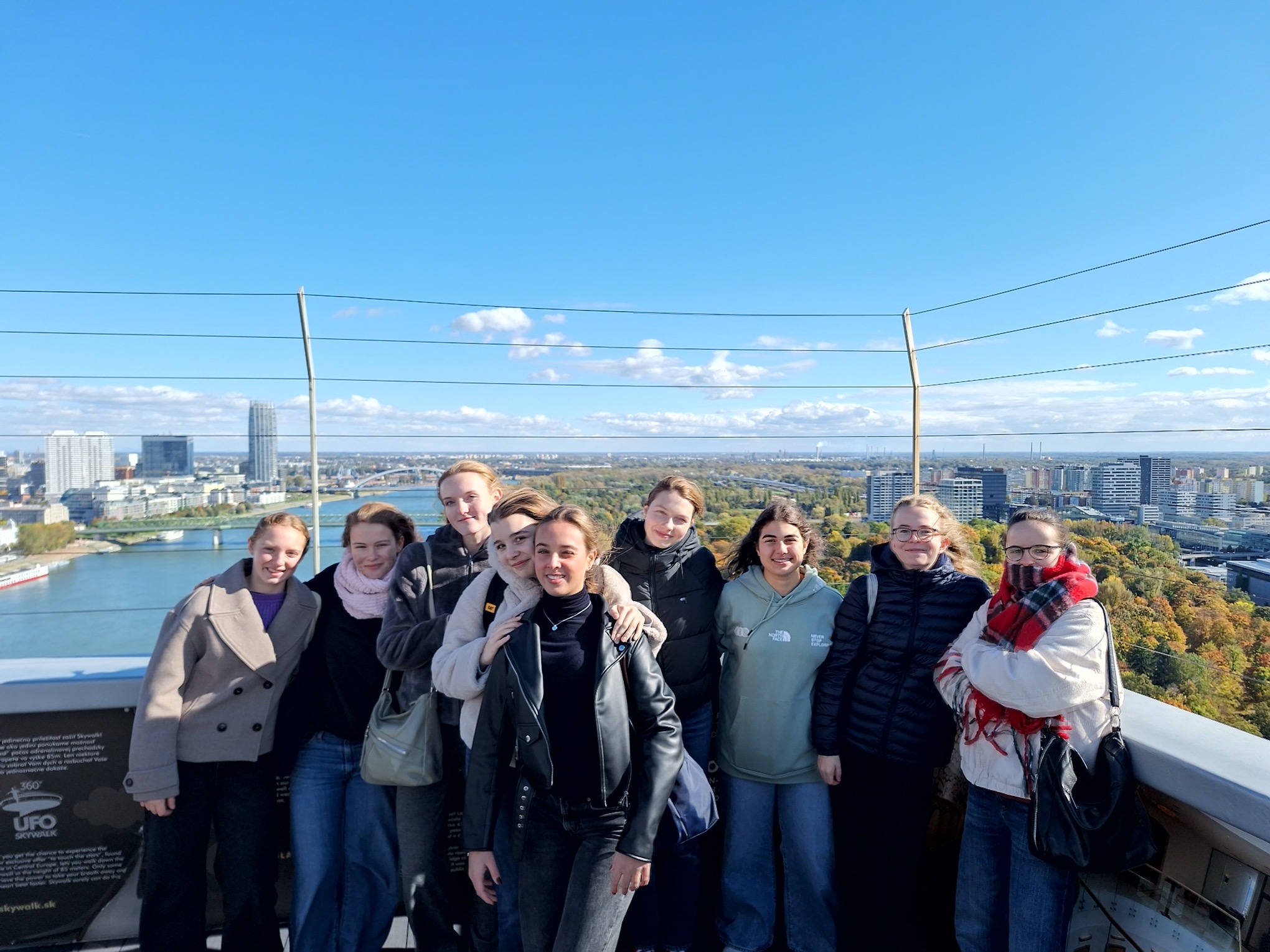 Gruppe von jungen Frauen in warmer Kleidung an einem sonnigen Tag auf einem Turm stehend mit einem Fluß und einer Stadt im Hintergrund.