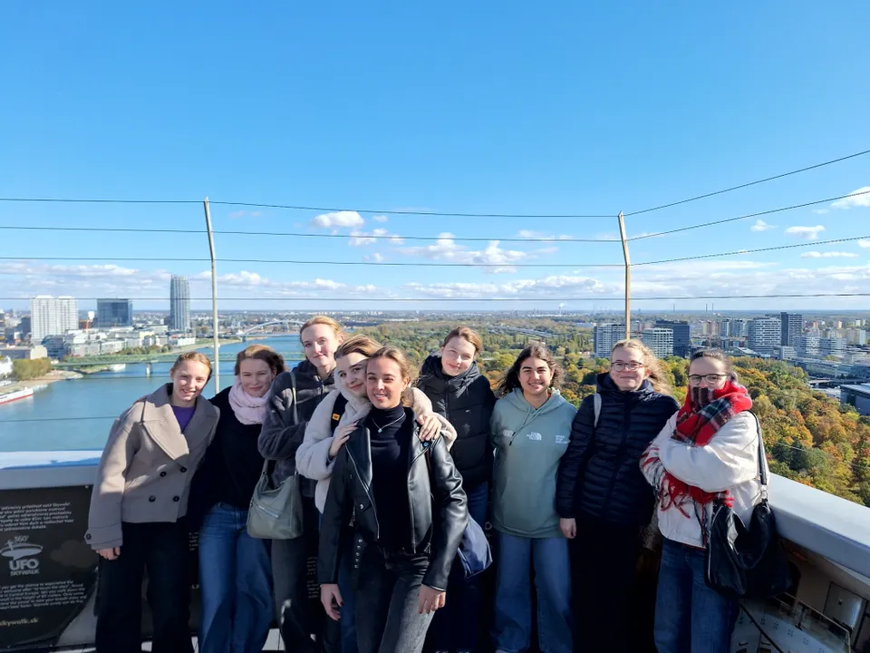 Gruppe von jungen Frauen in warmer Kleidung an einem sonnigen Tag auf einem Turm stehend mit einem Fluß und einer Stadt im Hintergrund.
