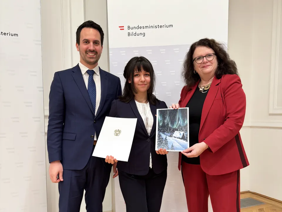 Zwei Frau und ein Mann in Geschäftskleidung vor einem Roll up des Bildungsministeriums mit der Frau in der Mitte eine Zeichnung und eine Auszeichnung in die Kamera haltend.