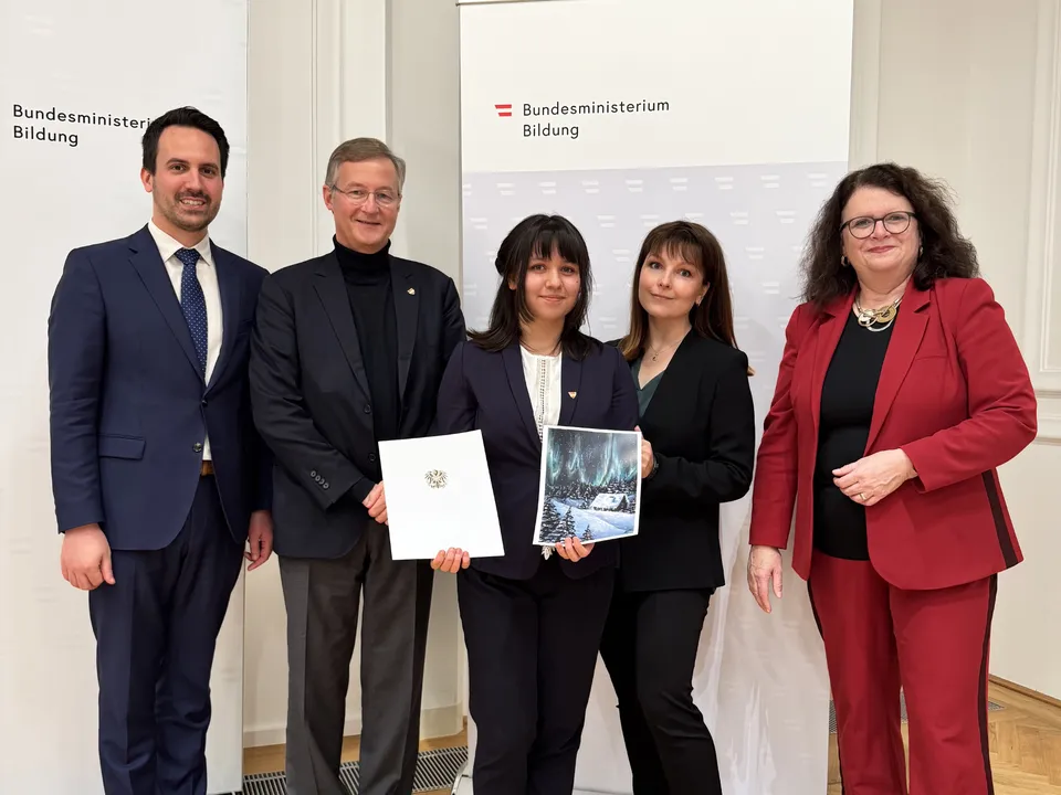 Gruppe von Erwachsenen in Geschäftskleidung vor einem Roll up des Bundesministeriums für Bildung und einer jungen Frau zwei Dokumente in die Kamera haltend