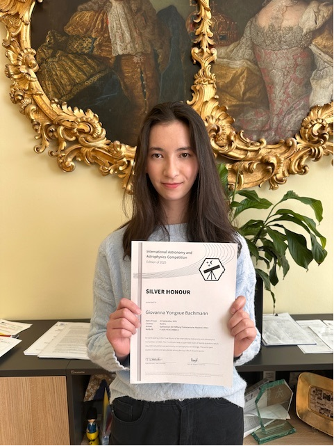 junge Frau mit langen schwarzen Haaren hält mit freundlichem Lächeln eine Urkunde in die Kamera