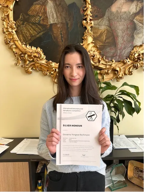 junge Frau mit dunklen langen Haaren hält eine Urkunde lächelnd in die Kamera