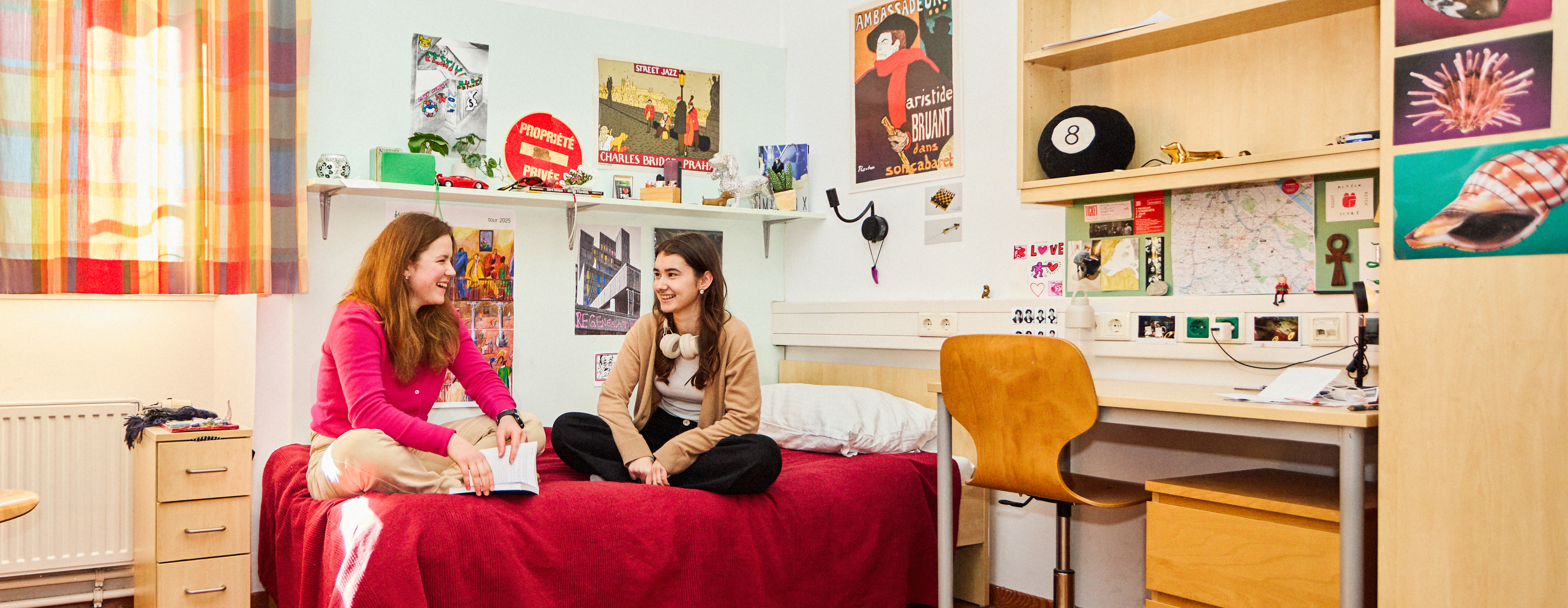 Zwei junge Mädchen in einem bunten Jugendzimmer auf einem Bett gemeinsam sitzend und zueinander gewandt und lachend.
