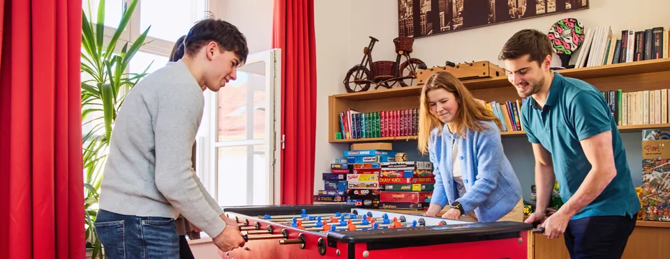 Zwei Burschen und ein Mädchen vertieft in ein Tischfussballspiel in einem Zimmer mit Bücherregal im Hintergrund, roten Vorhängen und mit offenem Fenster