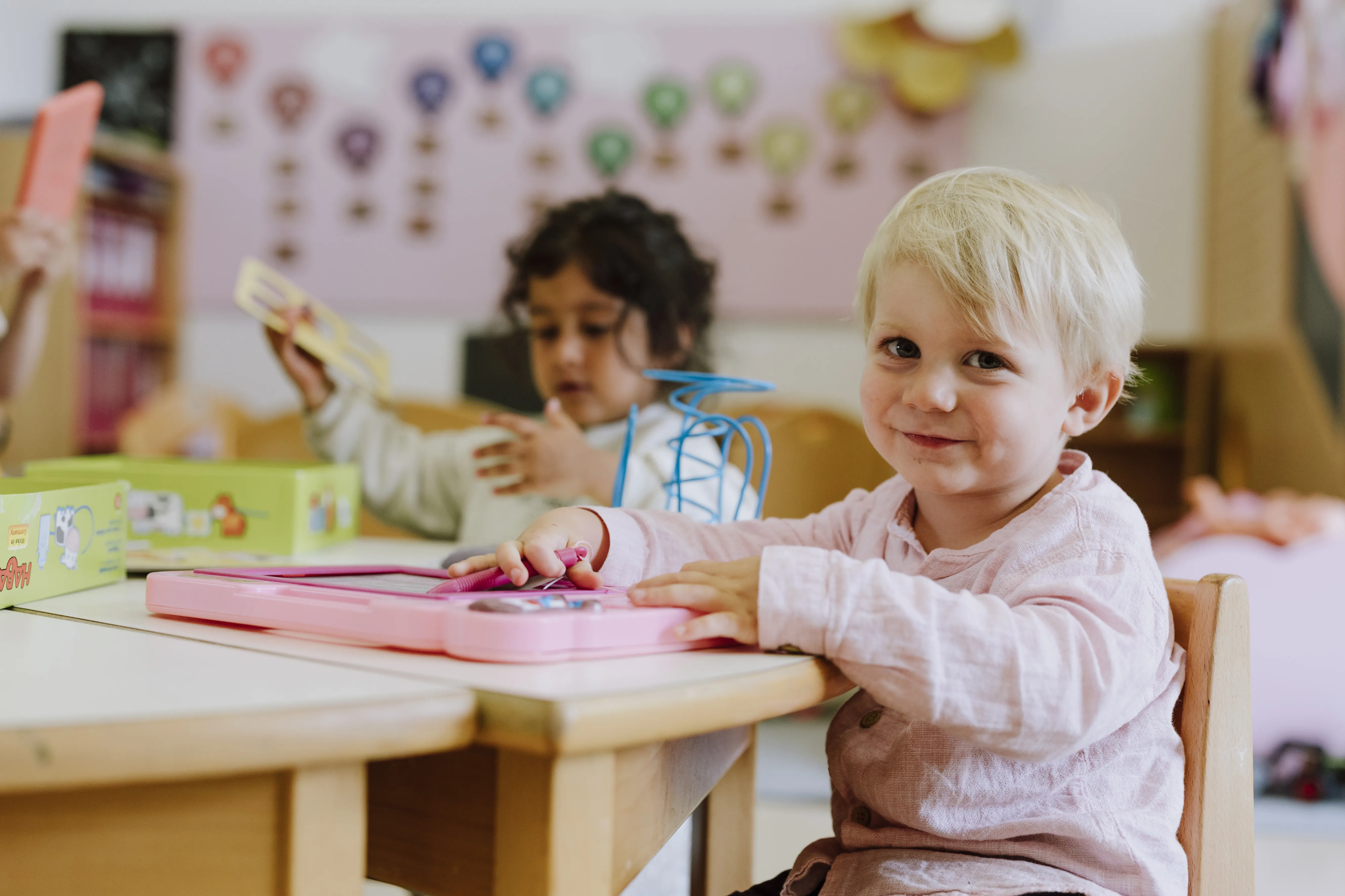 Lachende blondes kleines Kind spielend an einem Kindertisch sitzend mit einem Mädchen im Hintergrund.