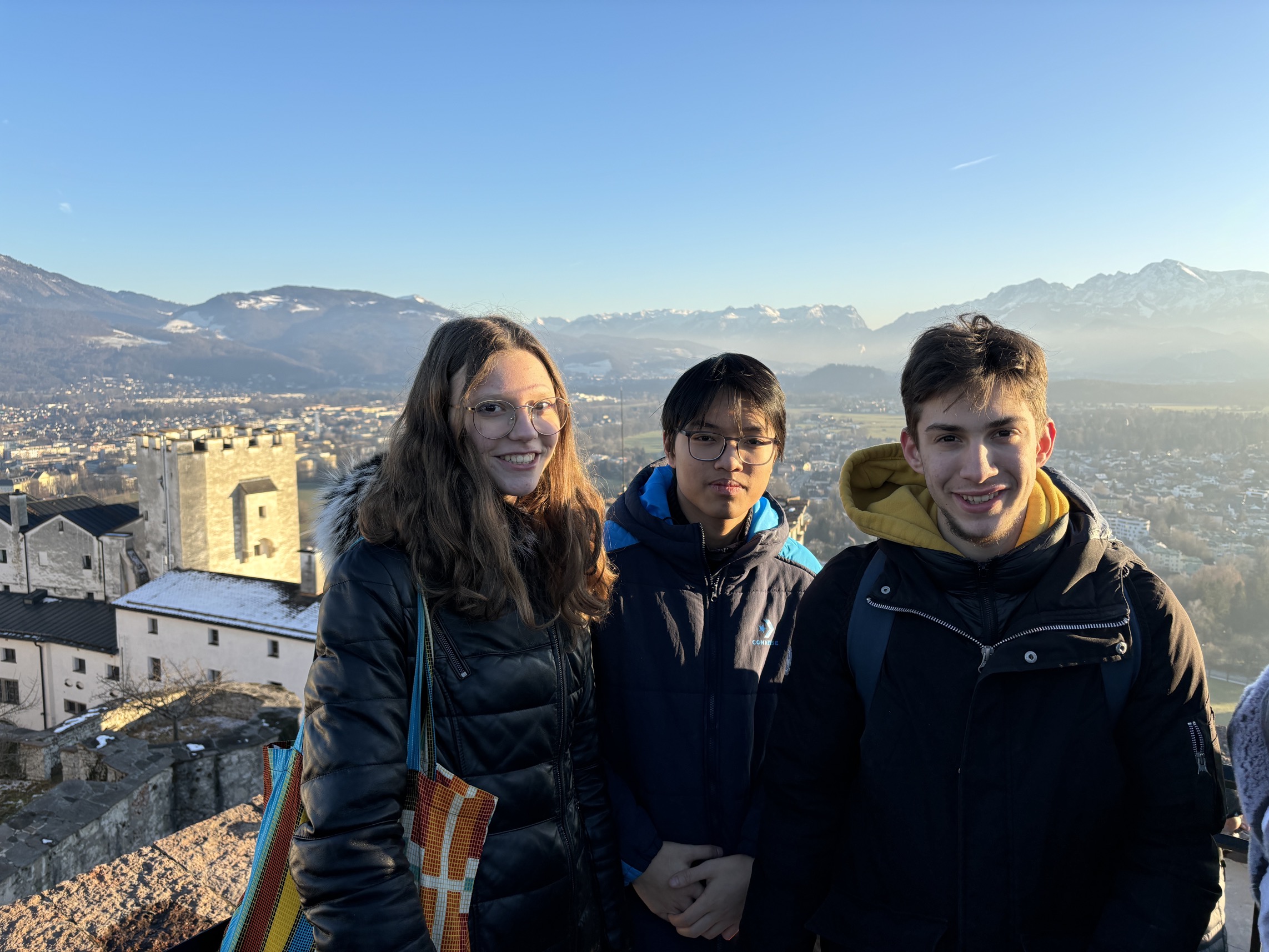 Eine junge Frau und zwei junge Männer in Winterkleidung bei sonnigem Wetter in die Kamera lachend mit einer Stadt und Landschaft im Hintergrund.