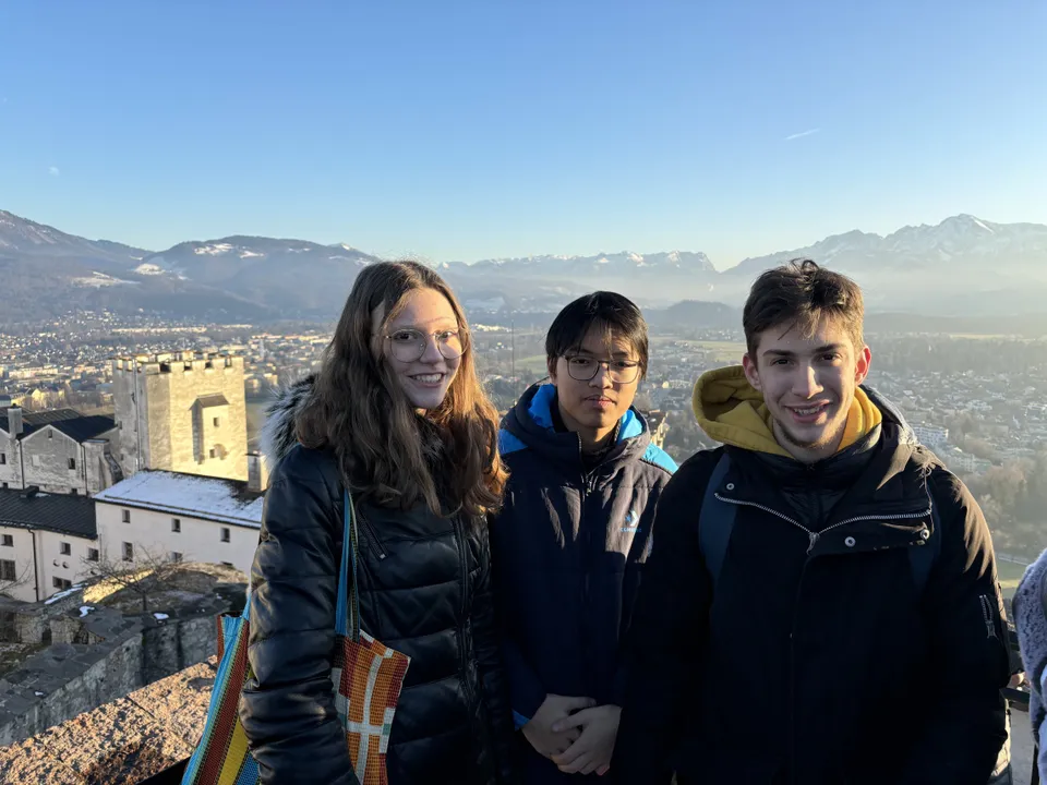 Eine junge Frau und zwei junge Männer in Winterkleidung bei sonnigem Wetter in die Kamera lachend mit einer Stadt und Landschaft im Hintergrund.
