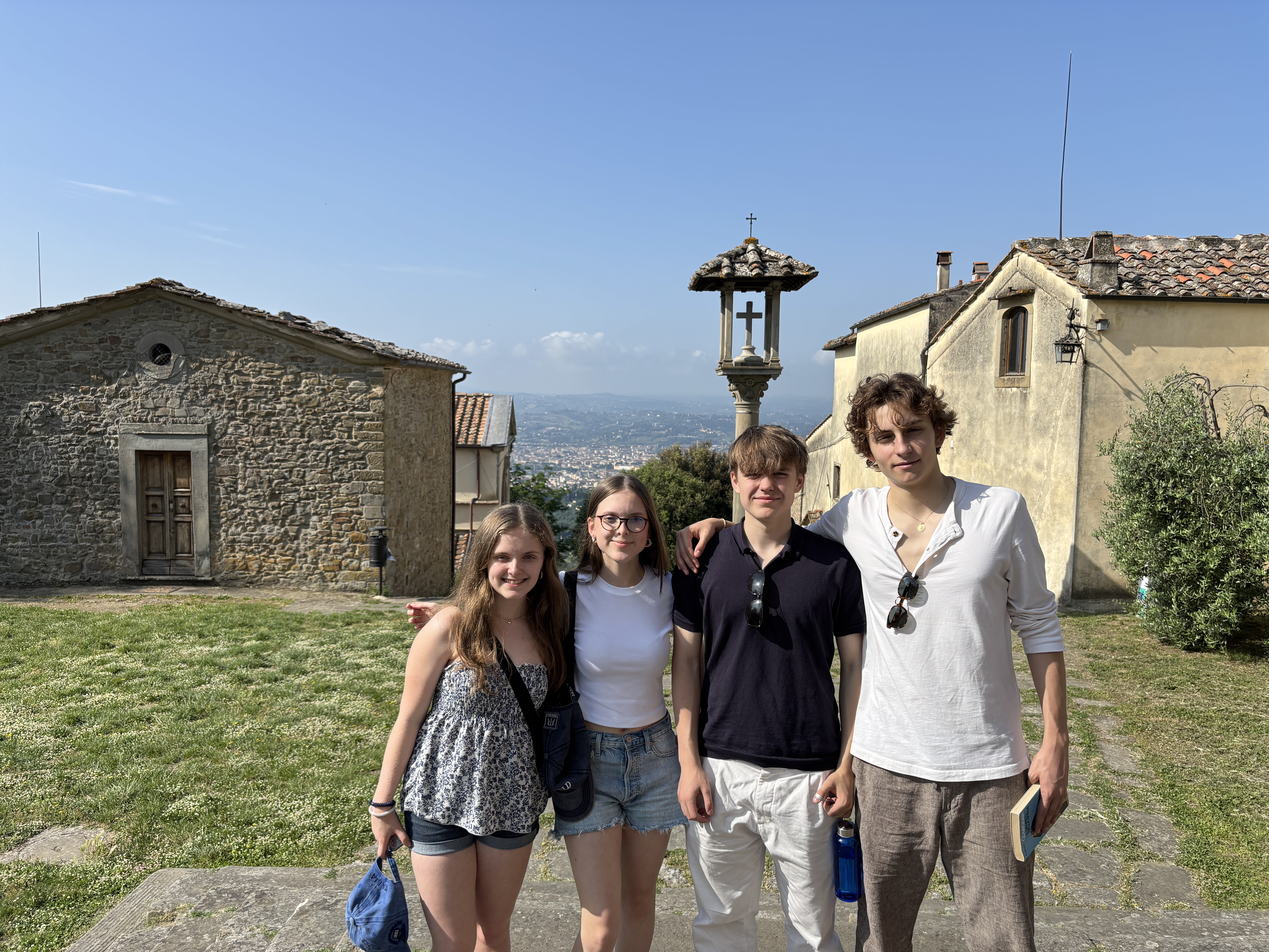 Gruppe von vier Jugendlichen in sommerlichen Outfits bei strahlend blauem Himmel sich umarmend und in Urlausstimmung in die Kamera lachend vor einem alten Steinhaus und Ausblick in das besiedelte Hinterland