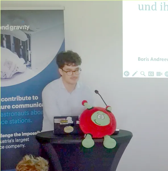 Junger Mann mit Brille und Oberlippenbart an einem Rednerpult stehend mit Projektion einer Präsentation im Hintergrund