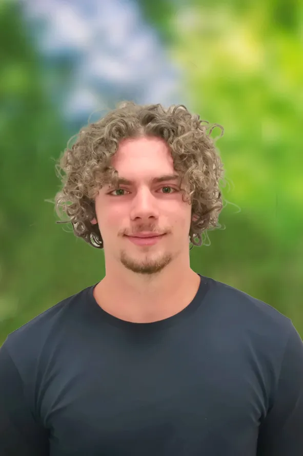 Portrait eines jungen Burschen mit lockigen Haaren und Bart mit blauen T-Shirt vor grünem verschwommenen Hintergrund