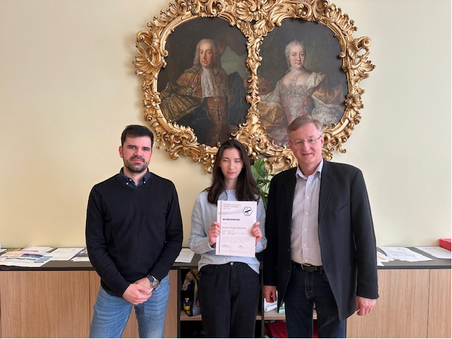 eine Schülerin mit einem Zertifikat in Händen haltend vor einem barocken Doppelportrait und links und rechts von männlichen Personen flankiert