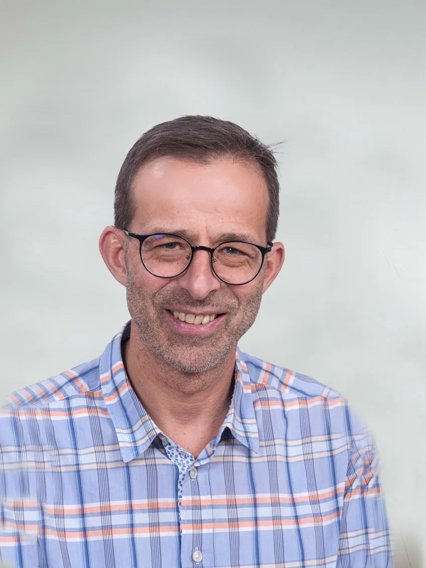 Portraitfoto eines sympathischer Mann mittleren Alters mit dunkel umrandeter Brille, 3-Tage-Bart und hellblau gemustertem Hemd vor neutral grauem Hintergrund
