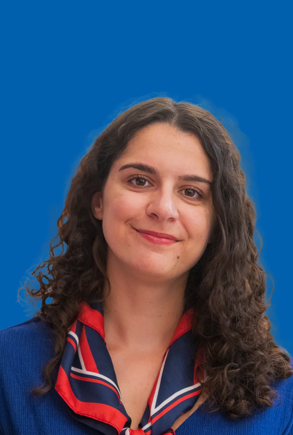 Junge Frau mit einem freundlichen Gesichtsausdruck, langem dunklem lockigen Haar und einem rot blauen Halstuch.