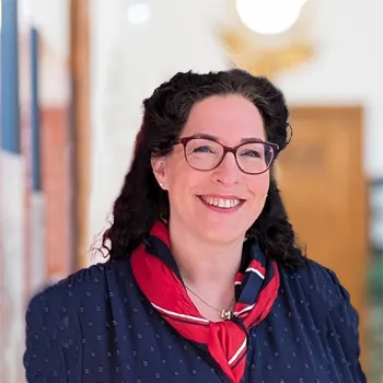 Frau mit Brille und dunklen Haaren, die lächelt und ein dunkelblau gemustertes Oberteil mit einem rot-blauen Tuch trägt.