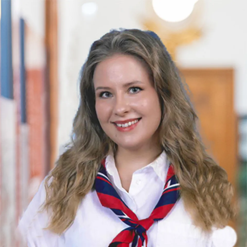 Portraitfoto einer freundlich lachenden jungen Frau mit langen, blonden welligen Haaren, mit weisser Bluse und dem offiziellen Halstuch des Theresianums. Blau, rot, weiss.