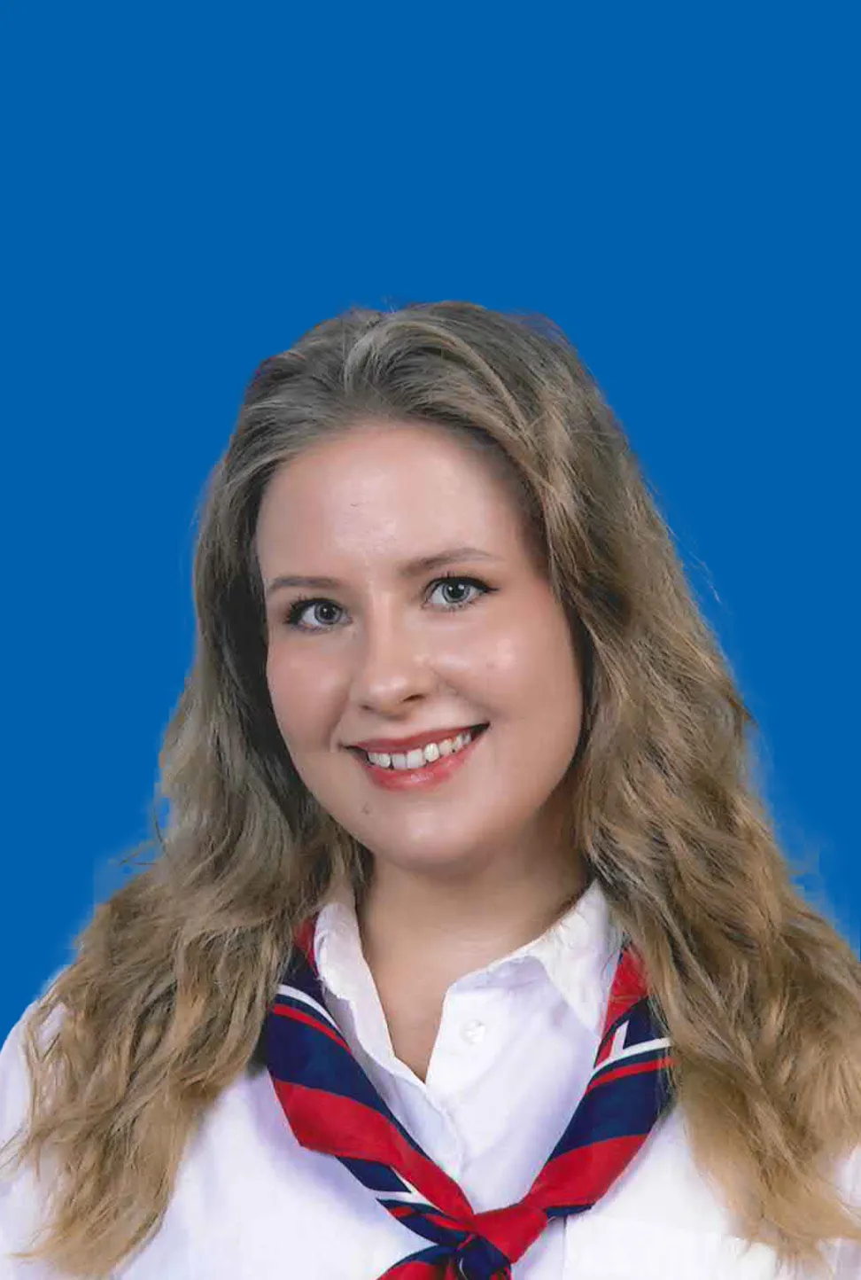 Portraitfoto einer freundlich lachenden jungen Frau mit langen, blonden welligen Haaren, mit weisser Bluse und dem offiziellen Halstuch des Theresianums. Blau, rot, weiss.