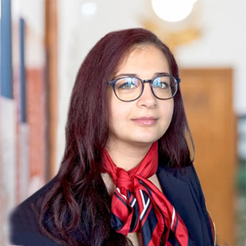 Eine junge Frau mit dunkelrotem, langem Haar, Brille und einem rot‑blauen Halstuch, die einen freundlichen Ausdruck zeigt.