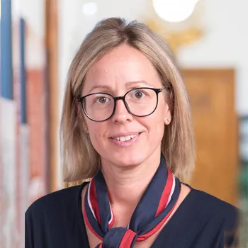 Frau mit kurzem blonden Haar, die eine Brille und ein rot blaues Halstuch trägt, die ein warmes Lächeln zeigt.