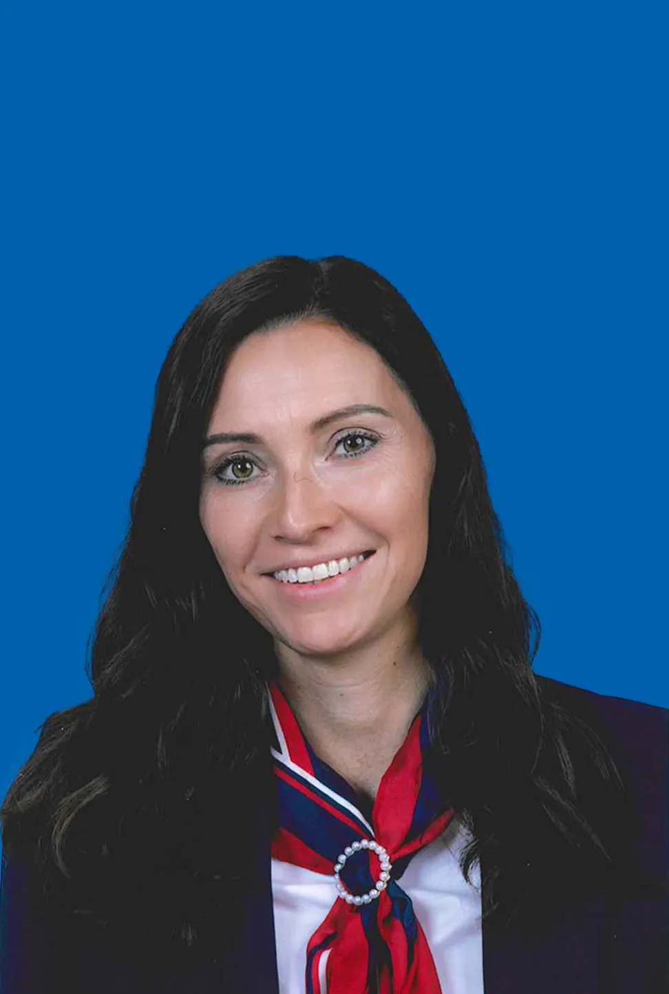 Lächelnde Frau mit langem dunklem Haar, die eine Jacke mit weißer Bluse und ein rot blaues Halstuch trägt.