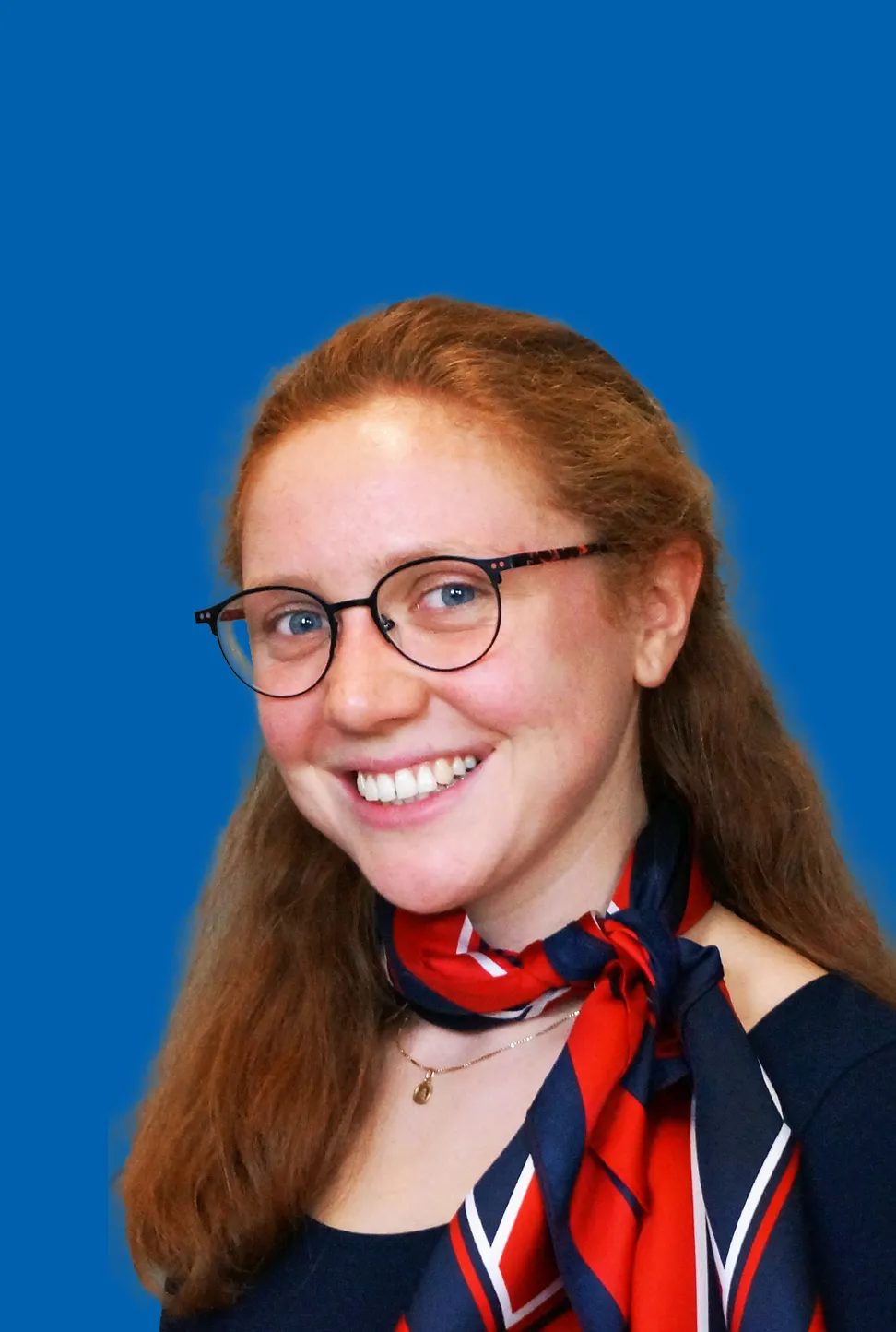 Lächelnde Frau mit roten, langen Haaren, einer Brille und einem seitlich gebundenen rot blauen Halstuch.