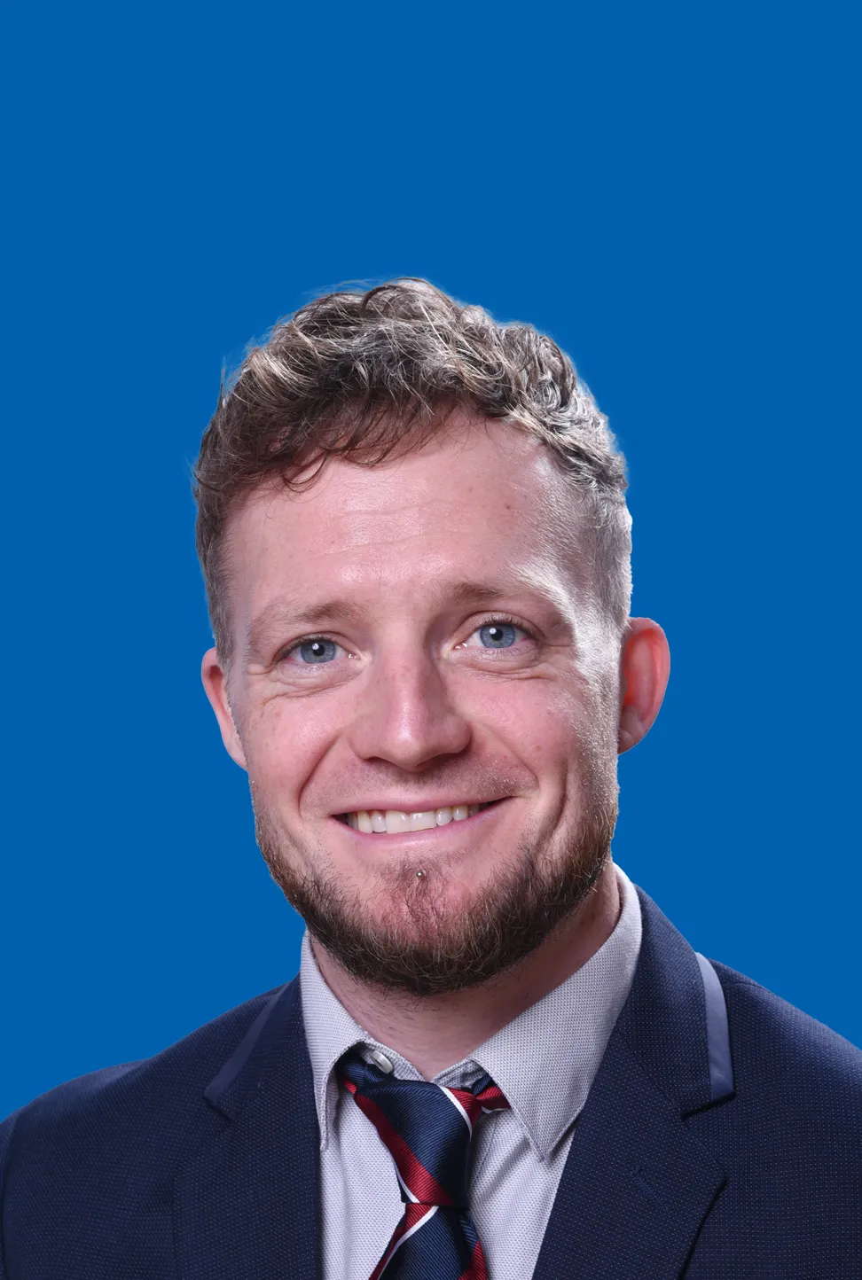 Mann mit rot-blondem Haar und Bart in einem dunklen Anzug und gestreifter Krawatte, der lächelt.