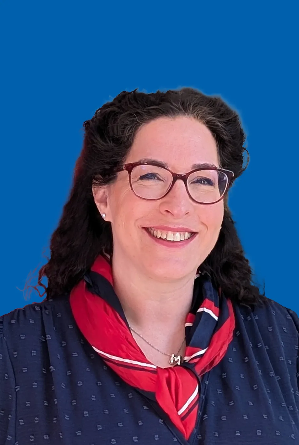 Frau mit Brille und dunklen Haaren, die lächelt und ein dunkelblau gemustertes Oberteil mit einem rot-blauen Tuch trägt.