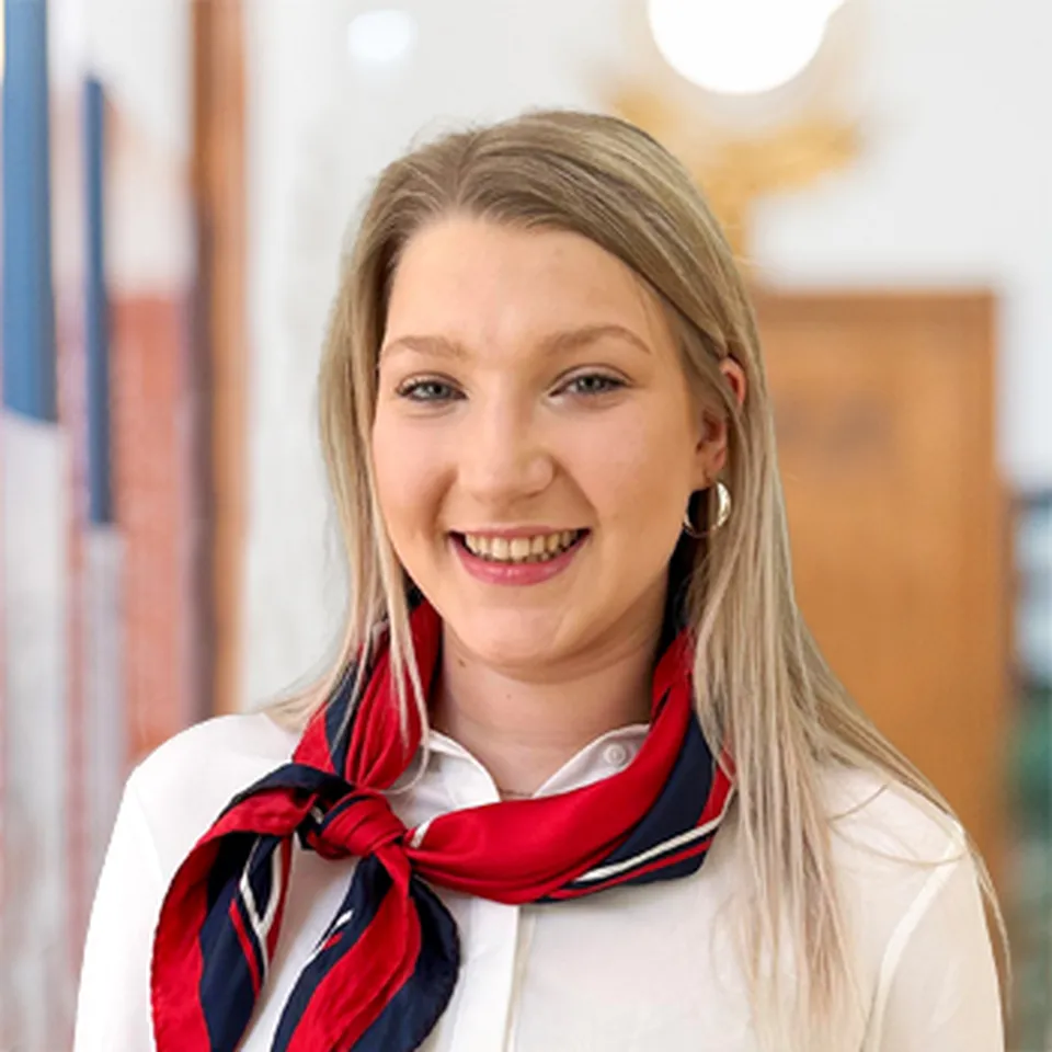 Eine junge Frau mit langem blondem Haar, die Ohrringe und ein rot blaues Halstuch trägt und freundlich lächelt.