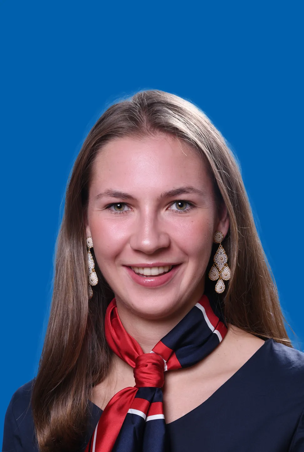 Junge Frau mit langen, dunkelblonden Haaren, die Ohrringe und ein rot‑blaues Halstuch trägt und ein freundliches Lächeln zeigt.