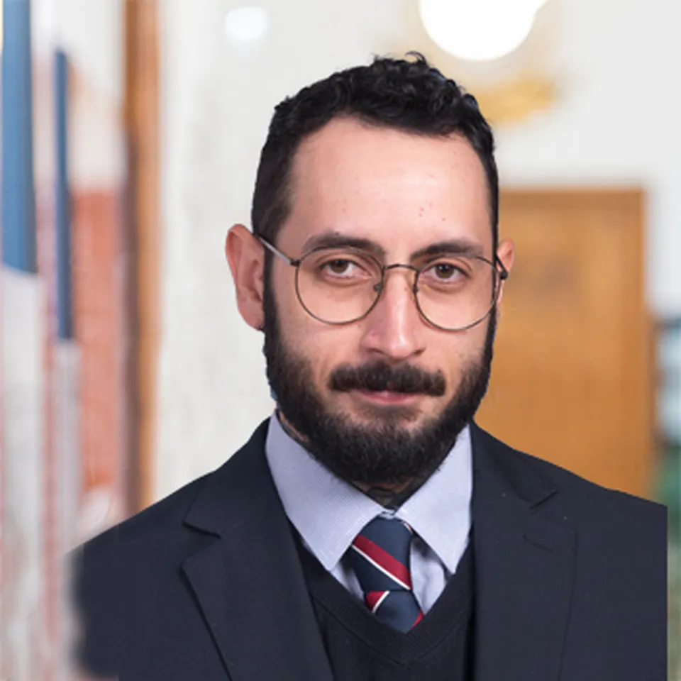 Mann mit dunklem Haar, Vollbart und Brille in einem dunklen Anzug und gestreifter Krawatte.