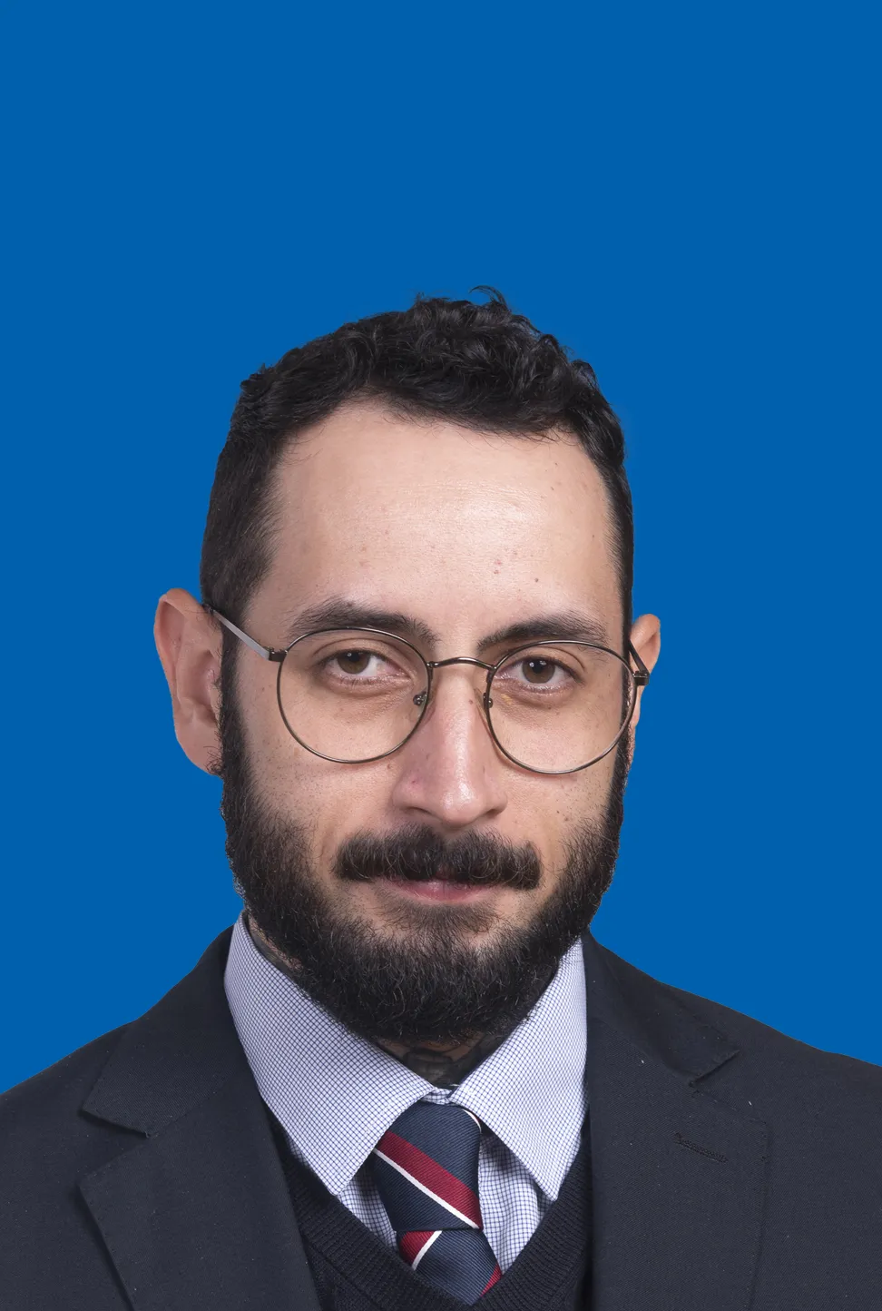 Mann mit dunklem Haar, Vollbart und Brille in einem dunklen Anzug und gestreifter Krawatte.