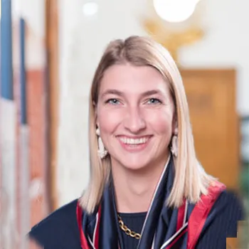 Blonde lächelnde Frau, die Ohrringe, Kette und ein rot-blaues Tuch trägt.