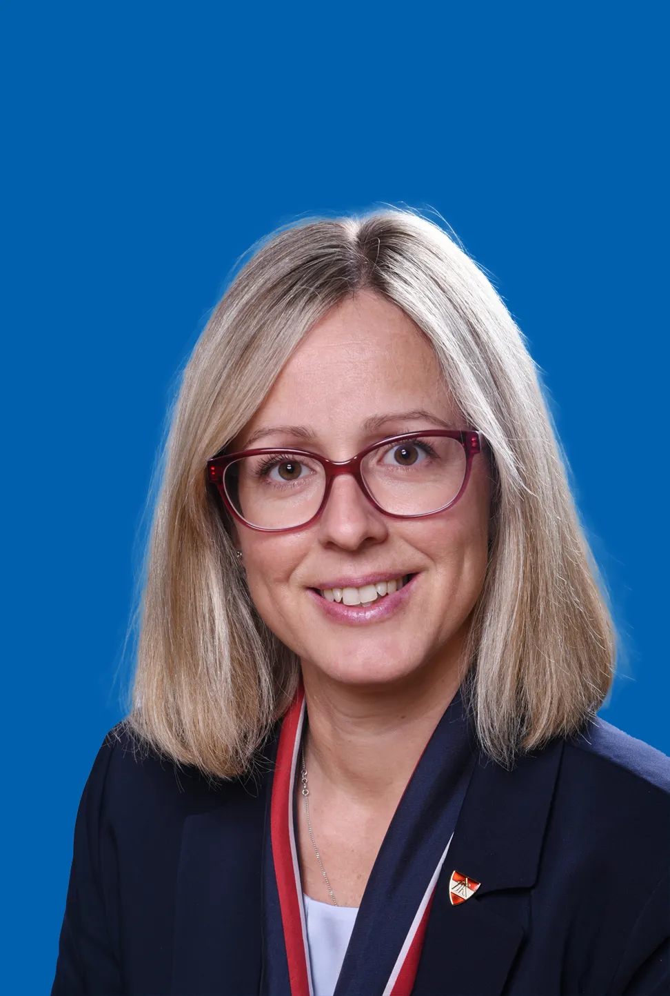 Frau mit kurzem blonden Haar, die eine Brille und ein rot blaues Halstuch trägt, die ein warmes Lächeln zeigt.