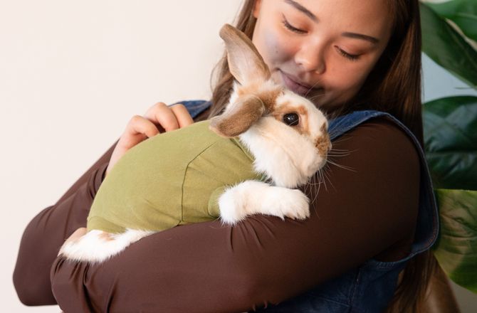 A woman sitting and cradling a rabbit wearing a green recovery suit, both appearing content and relaxed.