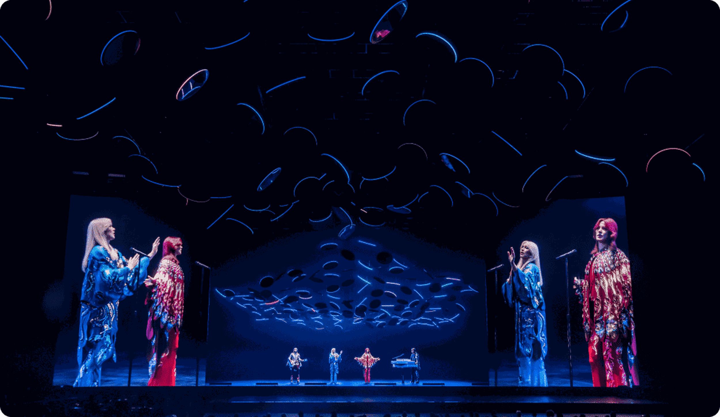 Concert stage with vibrant lighting and visuals. Four performers in colorful outfits sing and play instruments under illuminated circular fixtures.