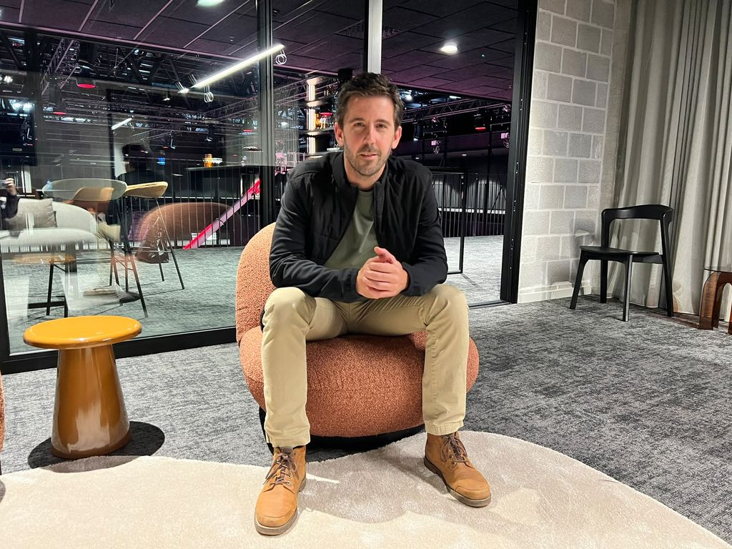 A man sitting on a round chair in a modern, well-lit interior with glass walls and stylish furniture, wearing a black jacket and beige pants.